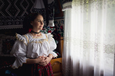 Young woman looking away while standing against window