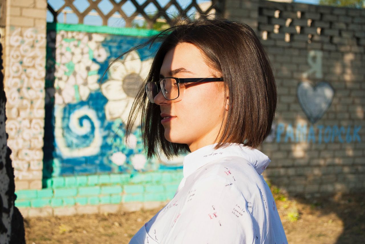 one person, headshot, real people, young adult, portrait, focus on foreground, lifestyles, leisure activity, hairstyle, side view, young women, glasses, looking, wall - building feature, eyeglasses, casual clothing, adult, graffiti, day, hair, beautiful woman, outdoors, profile view, brick