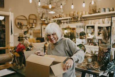 Happy senior female entrepreneur opening cardboard box at interior shop