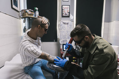 Side view of focused male making laser tattoo on arm of tired shaved client in tattoo salon