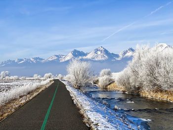 Winter landscape of bike path next to river in frozen field and snowy mountains in background