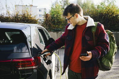 Man holding smart phone while charging electric car at station