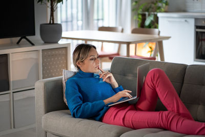 Focused attractive caucasian woman writer with laptop writes book lies on sofa in living room