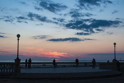 Silhouette people on street by sea against sky during sunset