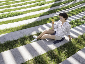 Woman texting in her smartphone while sits on bench in urban park. striped shadow on green lawn. 