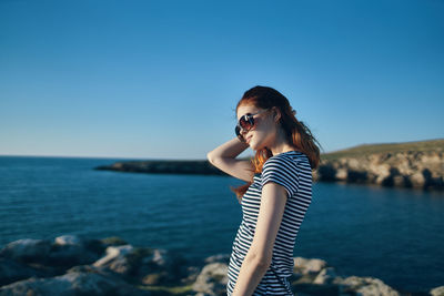 Young woman wearing sunglasses standing by sea against clear blue sky