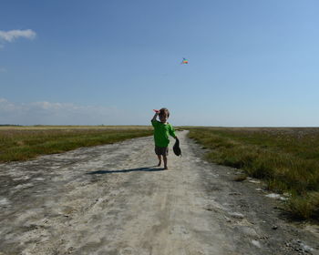 Boy flying kite while standing on road amidst grassy field against sky during sunny day