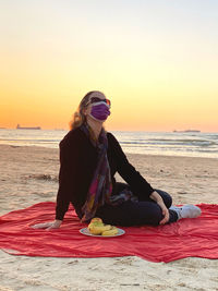 Woman wearing mask sitting on blanket at beach during sunset