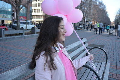 Happy woman holding pink balloons while sitting on bench in city