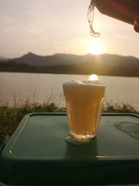 Close-up of drink by lake against sky during sunset