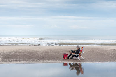 Mature adult male sitting on a beach chair pouring water in a mate and behind the waves of the sea.