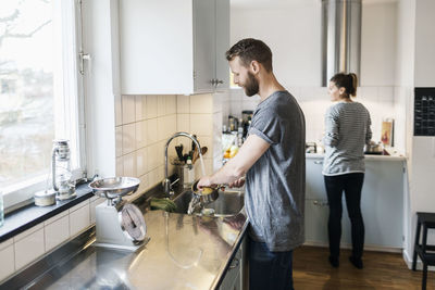 Man washing sauce pan while woman standing in background in kitchen