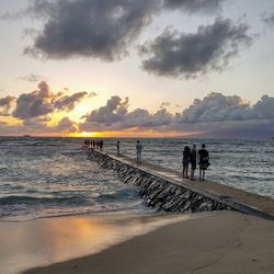 People standing on pier at beach against cloudy sky during sunset