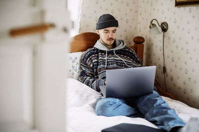 Young man using laptop while relaxing on bed seen through doorway at home