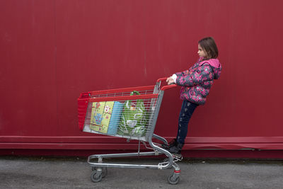 Full length of girl standing on shopping cart against red wall