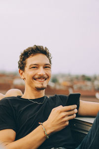 Portrait of smiling young man using mobile phone while sitting on terrace against sky