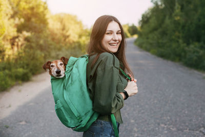 A smiling girl is holding a green backpack on her shoulder, from which a cute dog looks out