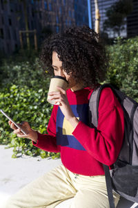 Young woman drinking coffee while using phone while sitting at park