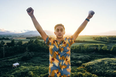 Portrait of young man with arms raised standing against sky
