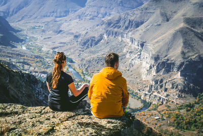 Rear view of couple sitting on cliff against mountains