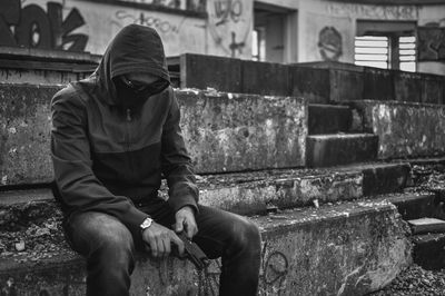 Hooded man holding handgun while sitting on steps against abandoned building