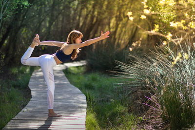 Full length of woman doing yoga on boardwalk against plants