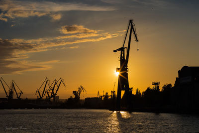 Silhouette of cranes at harbor against sky during sunset