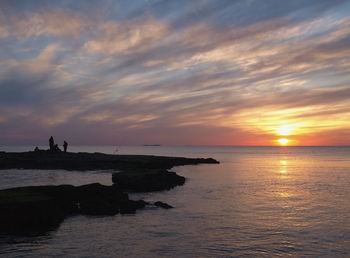 Silhouette people on cliff by sea against sky during sunset