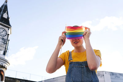 Smiling non-binary person covering face with rainbow flag