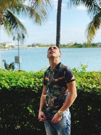 Young man looking up while standing against plants and lake