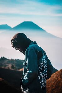 Young man wearing hooded shirt while standing against mountains and sky