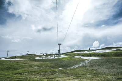 Low angle view of overhead cable car on grassy hill against cloudy sky