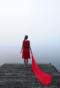 Rear view of woman holding scarf while standing on wooden pier by lake in foggy weather
