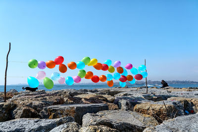Multi colored balloons on rock by sea against clear blue sky