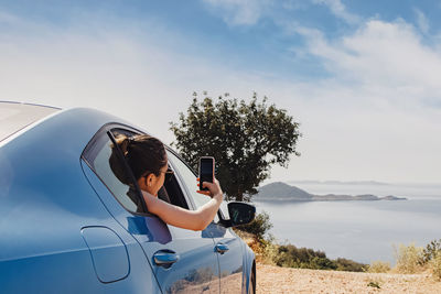 Young woman taking picture with smartphone from open window of a car stopped roadside with a view