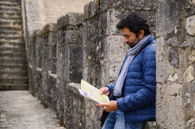 A man wearing a blue jacket studies a map standing against a fortress wall.