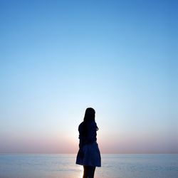 Rear view of woman standing by sea against clear sky during sunset