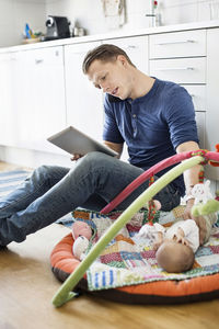 Father using digital tablet and mobile phone while baby sleeping on mat in kitchen