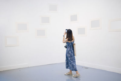 Woman holding camera while standing against white wall