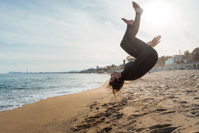 Full body side view of active young female in wetsuit performing acrobatic somersault in air while exercising on sandy beach near ocean