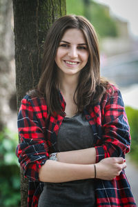 Portrait of smiling young woman standing against tree trunk