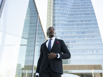 Serious african american male entrepreneur wearing elegant suit walking along glass office building in city center and looking away