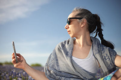 Portrait of young woman holding sunglasses against sky
