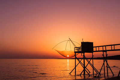 Silhouette fishing hut by sea against orange sky