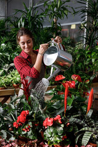 Adult woman in apron pouring clean water on green tropical plants while working in professional greenhouse