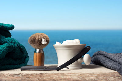 Close-up of razor with shaving cream and brush on table by sea against sky