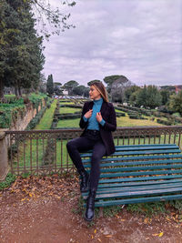 Full length portrait of woman sitting on bench against sky