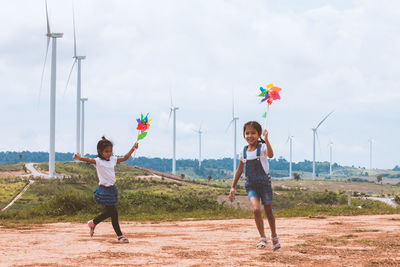 Full length of sisters playing with pinwheel toys on land with windmills in background