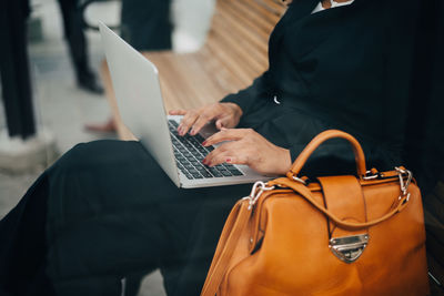 Midsection of businesswoman typing on laptop sitting at bus shelter seen from glass