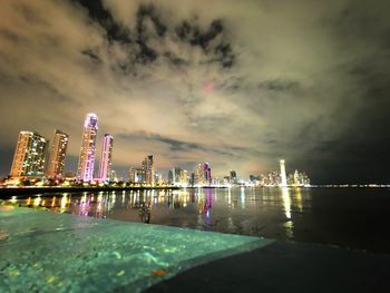Illuminated buildings by sea against sky at night